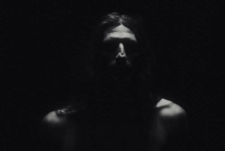 A shirtless man with long hair appears in flickering, glitchy flashes of light, looking terrified in a dark room