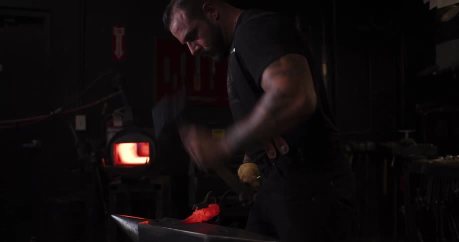 A strong blacksmith with tattoos hammers a piece of glowing red-hot metal on an anvil in his dark workshop