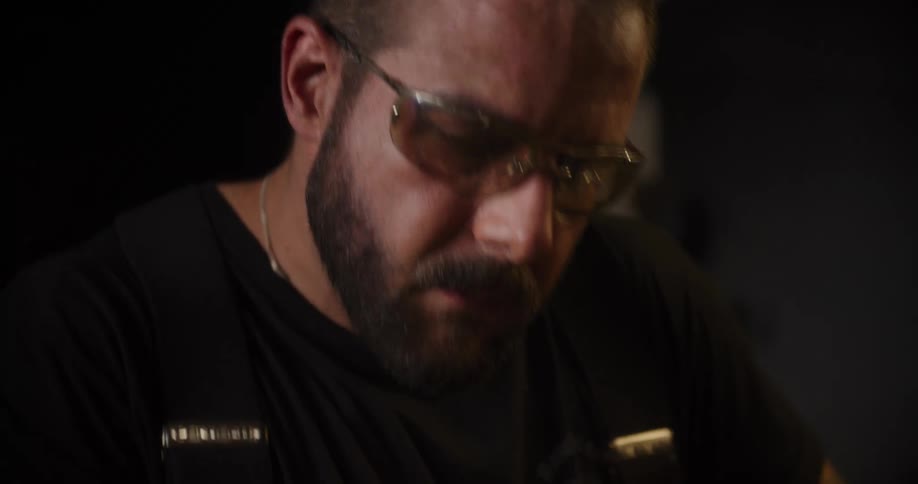 Close-up rack focus shot of a bearded craftsman in safety glasses concentrating on his work in a dimly lit workshop