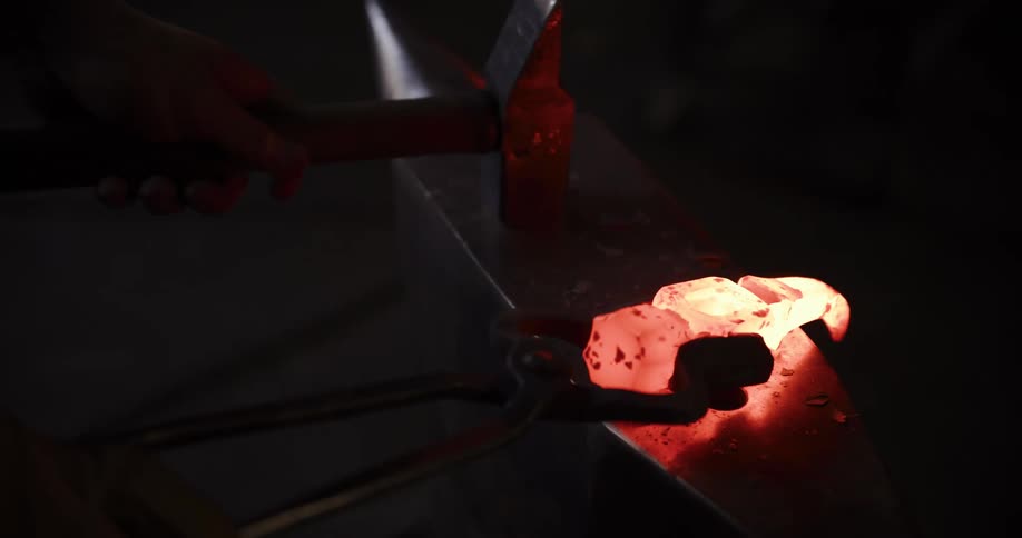 A blacksmith forges a glowing piece of hot metal with a hammer on an anvil in slow motion, sending sparks flying in a dark workshop
