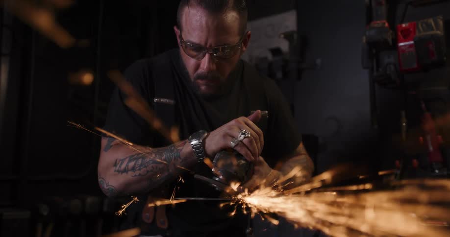 A focused metalworker with tattoos grinds a piece of steel, sending a brilliant shower of sparks flying in his workshop