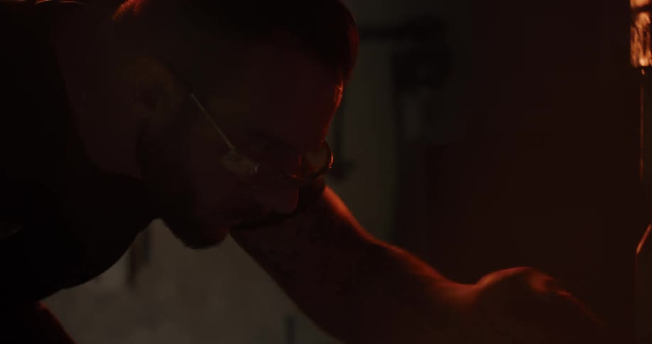 A focused blacksmith with safety glasses works intently in his dimly lit workshop, illuminated by the warm glow of the forge in slow motion