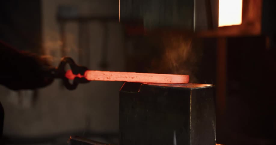 An industrial power hammer repeatedly strikes and flattens a glowing hot piece of metal held by tongs in a dark workshop
