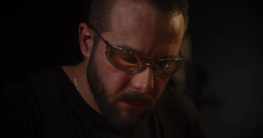Intense close-up of a bearded craftsman in safety glasses, concentrating on his work as flames flicker in the foreground
