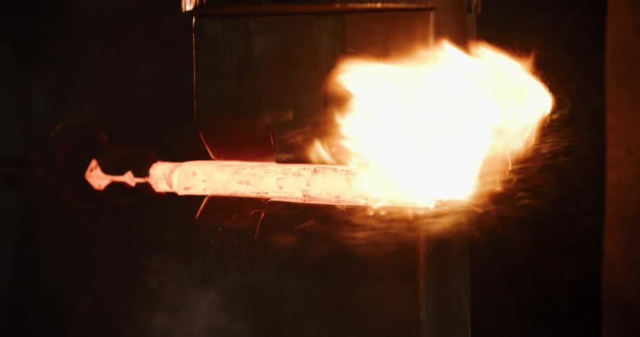 An industrial press hammers a glowing hot metal billet, shaping it with explosive force and sending sparks flying in a dark factory