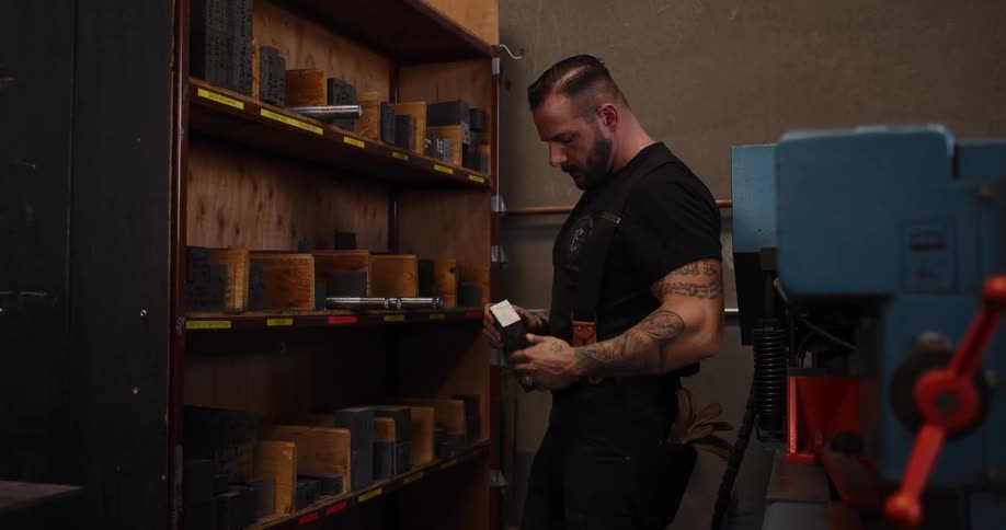 A muscular, tattooed worker in a workshop selects a metal block from a storage shelf