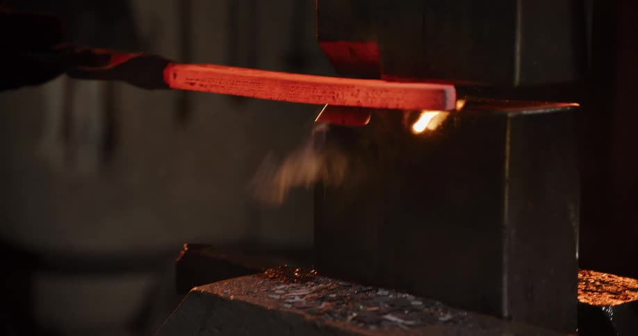 An industrial power hammer repeatedly strikes and flattens a glowing red-hot metal bar in a dark forge in slow motion