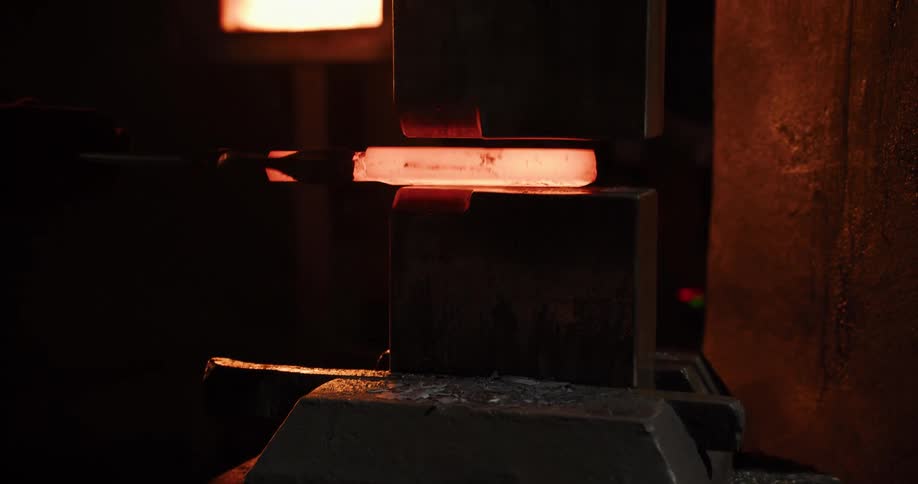 Close up of a powerful industrial press forging and shaping a piece of red-hot metal in a dark workshop