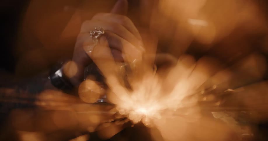 An artistic close-up with motion blur captures a metalworker's tattooed hands using an angle grinder, creating a fiery spray of sparks