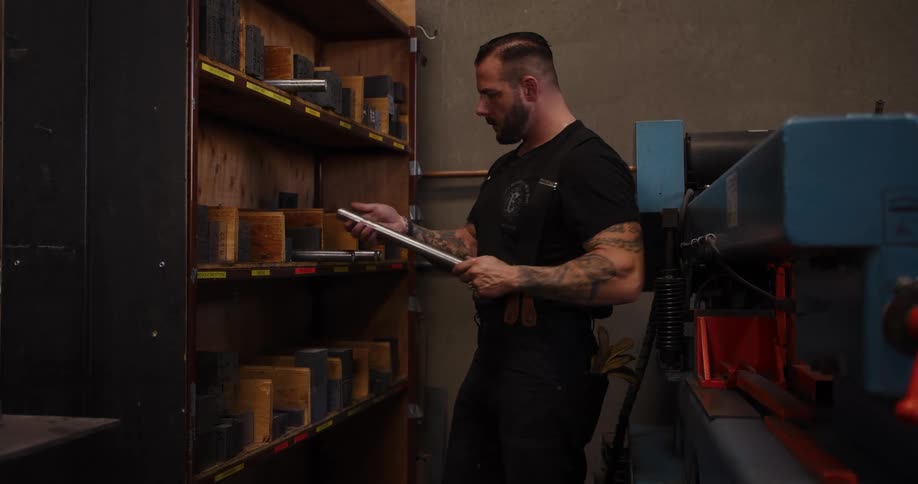 A tattooed metalworker carefully places a metal tool on a storage shelf in his workshop