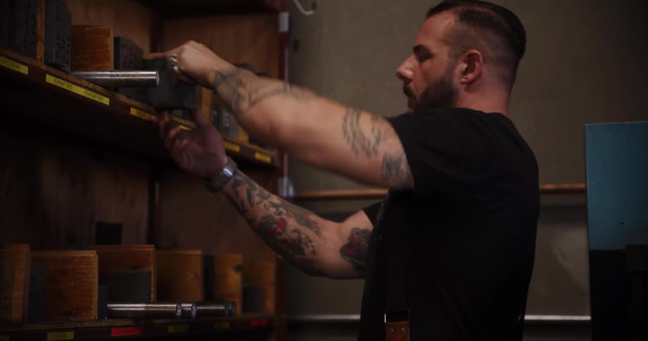 A muscular craftsman with tattoos on his arms carefully selects a metal block from a shelf in his workshop