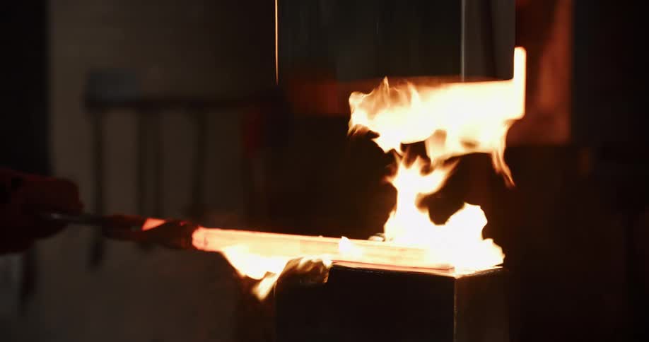 In a dimly lit forge, a blacksmith uses a powerful industrial hammer to shape a piece of glowing hot, flaming metal