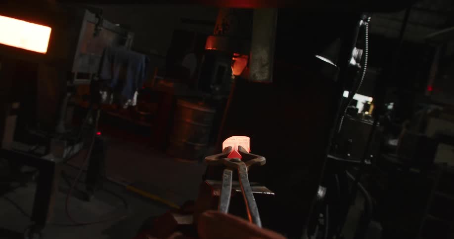 A metalworker uses tongs to pull a piece of glowing hot metal out of a furnace in a dark workshop
