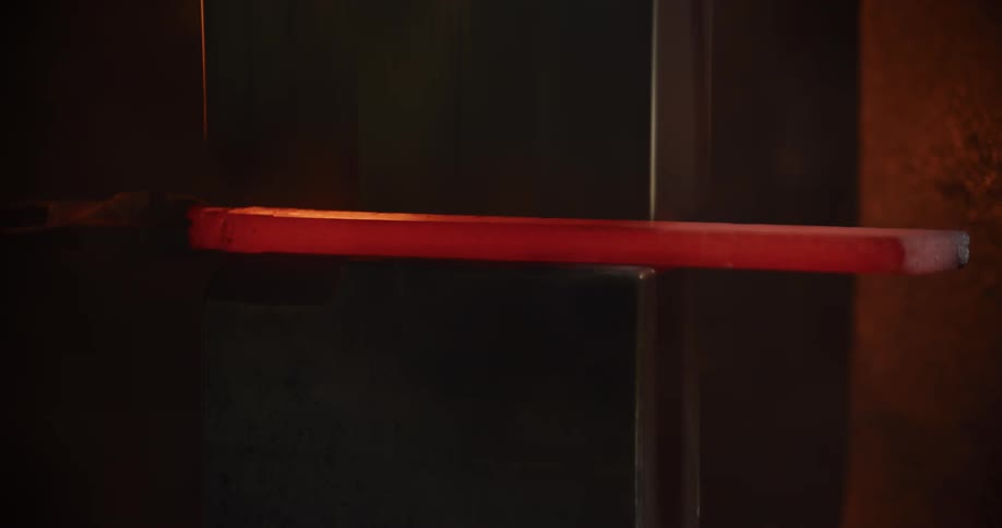 A massive industrial power hammer forges and shapes a glowing red-hot metal bar in a dark, smoky workshop
