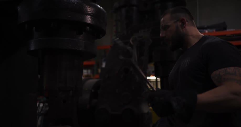A tattooed craftsman wearing safety glasses operates a large industrial power hammer in a dimly lit workshop before moving out of frame
