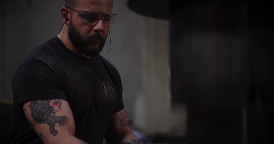 Close up of a focused blacksmith with tattoos and safety glasses working in his dimly lit forge