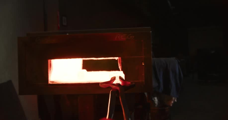 A glowing hot piece of metal is pulled from a furnace with tongs and then hammered by a powerful industrial press, causing flames to erupt