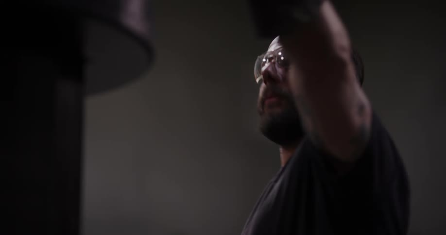 A focused industrial worker with a beard and safety glasses operates machinery in a dimly lit workshop