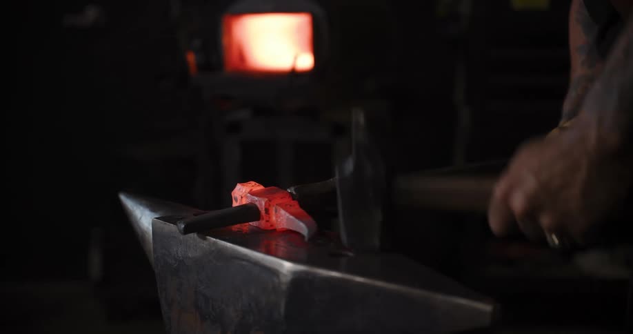A skilled blacksmith in a dark workshop hammers and shapes a glowing hot piece of metal on an anvil