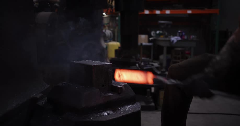 A blacksmith uses a power hammer to shape a glowing piece of hot metal and then returns it to a fiery furnace in slow motion
