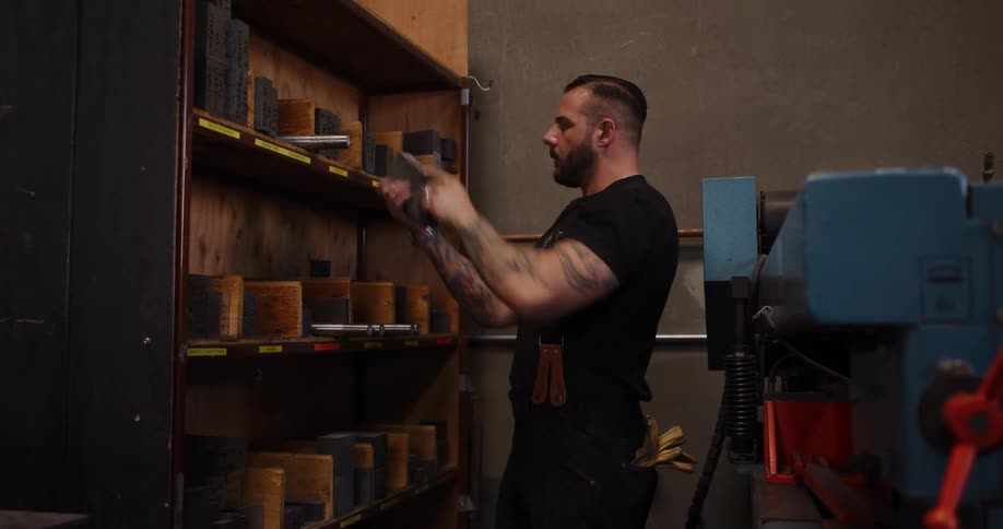 A muscular metalworker with tattoos selects tools and materials from a storage rack in his industrial workshop