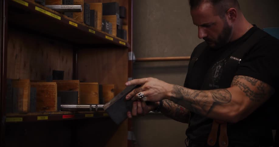 A tattooed artisan carefully selects a metal letterpress block from a wooden shelf in his workshop