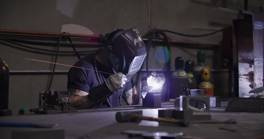 A tattooed welder in a protective mask TIG welds a metal piece on a workbench as the camera slowly pulls back