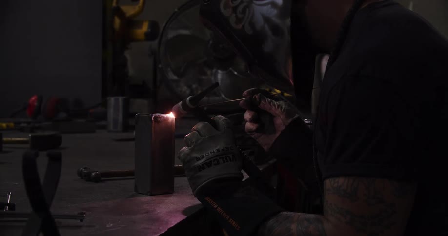 A craftsman in a dark workshop meticulously uses a TIG welder to fuse a metal block, creating a bright arc of light