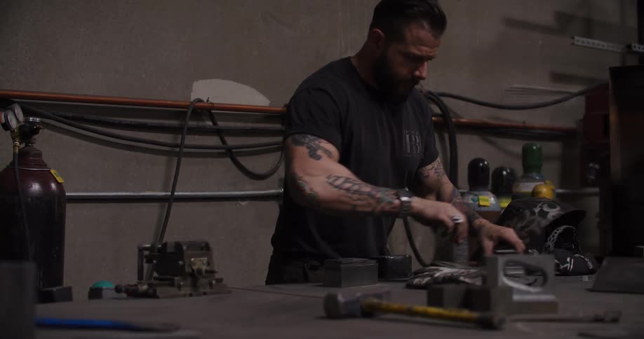 A muscular, tattooed metalworker prepares his welding station and tools in a dimly lit industrial workshop