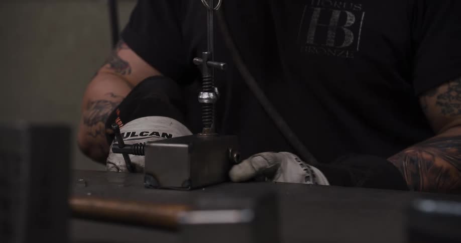 A tattooed metalworker in his workshop carefully adjusts a complex metal clamp on his workbench before beginning his work
