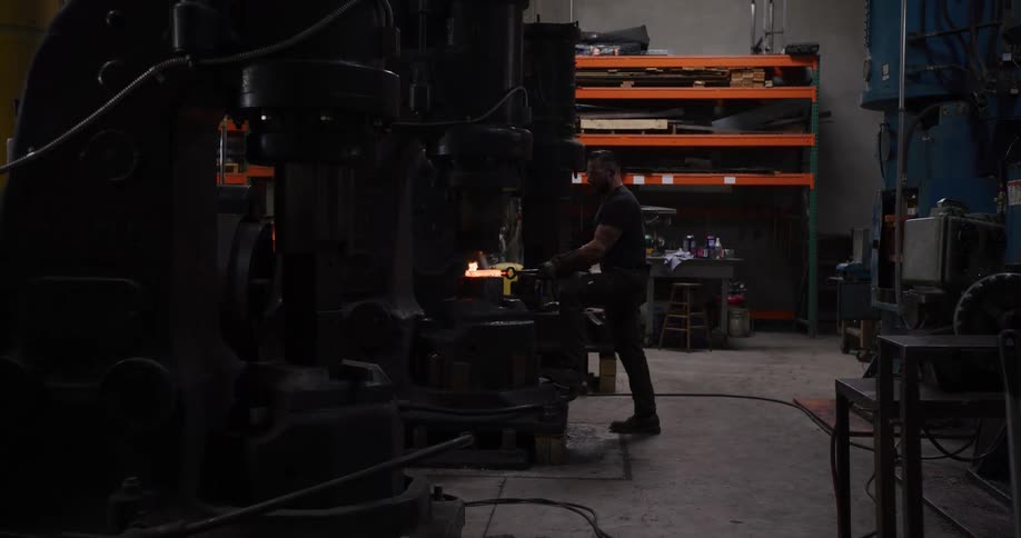 A blacksmith uses a large power hammer to forge a glowing hot piece of steel in a dimly lit industrial workshop