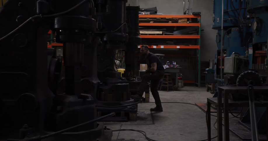 A blacksmith walks through his industrial workshop to operate a large power hammer, shaping metal with powerful blows