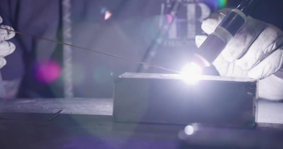 Slow motion tilt down from a welder's helmet to the bright arc of a TIG torch meticulously welding metal in a workshop