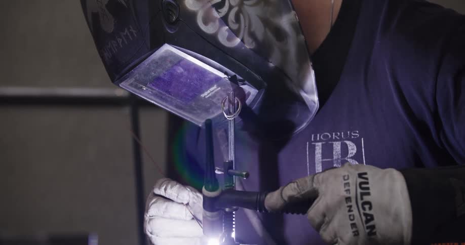 A skilled craftsman puts on his custom-painted helmet and carefully welds a small, intricate metal piece in his workshop