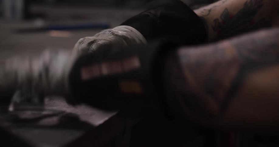A close-up of a tattooed craftsman wearing welding gloves meticulously clamping, assembling, and hammering metal parts together in a workshop