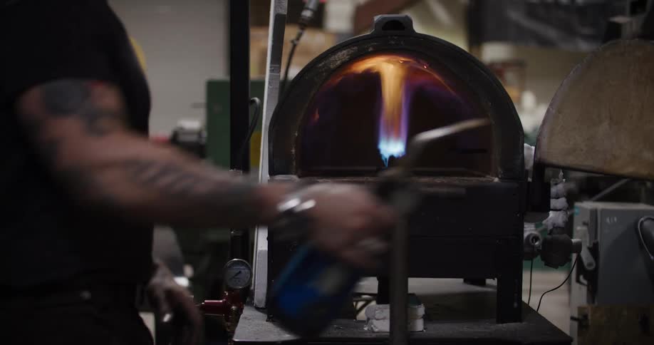 A craftsman with tattoos lights a large industrial forge with a propane torch in a workshop