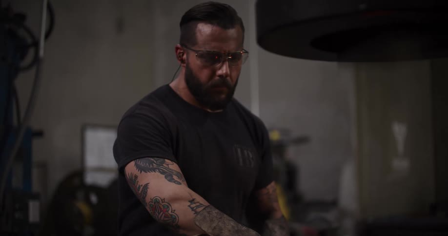 A focused blacksmith with tattoos and safety glasses operates a power hammer in his workshop