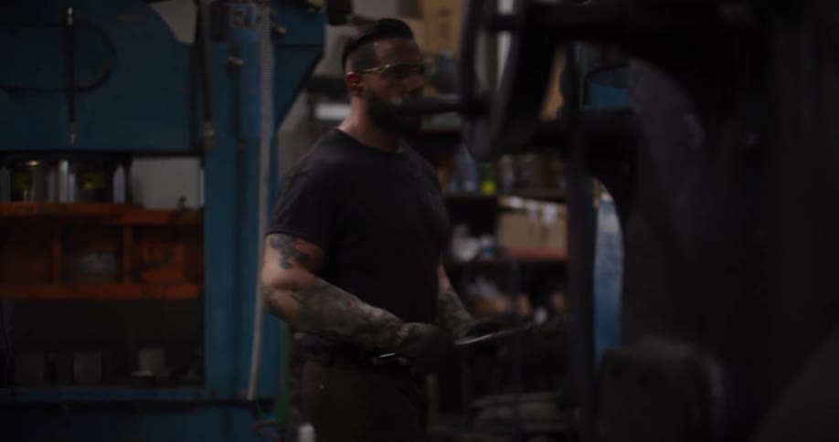 A tattooed male worker in a dark workshop turns off a large industrial machine and walks away with a tool
