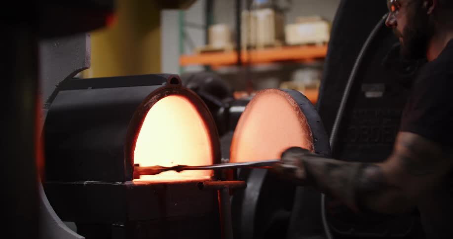 A tattooed blacksmith in a workshop uses tongs to carefully handle a glowing hot piece of metal in an industrial forge