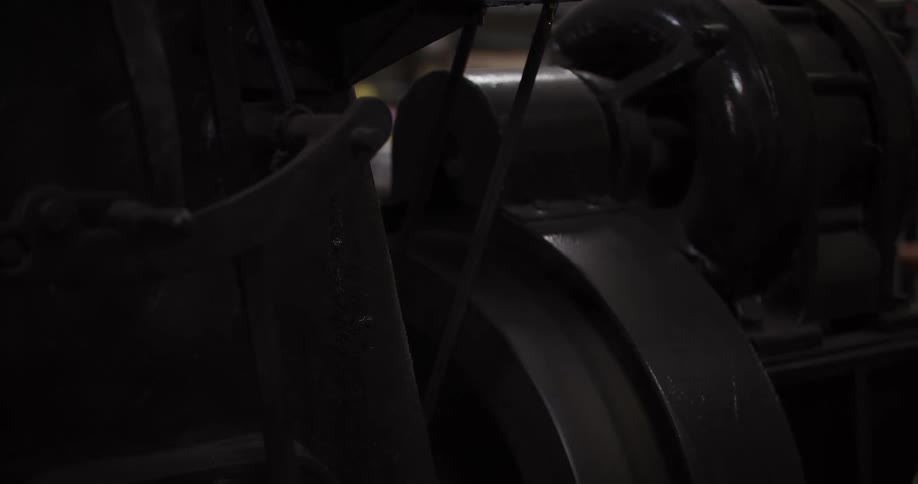 A close-up view of a large vintage industrial machine with a spinning wheel and belt in a dark factory setting