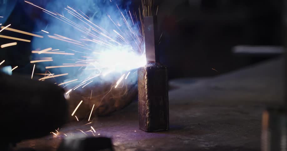 A skilled metalworker in a protective mask welds a metal joint, sending a brilliant shower of sparks flying in slow motion