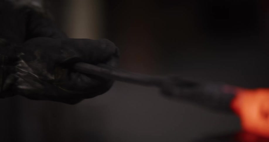 A blacksmith in a dark workshop uses tongs to hold a red-hot piece of metal