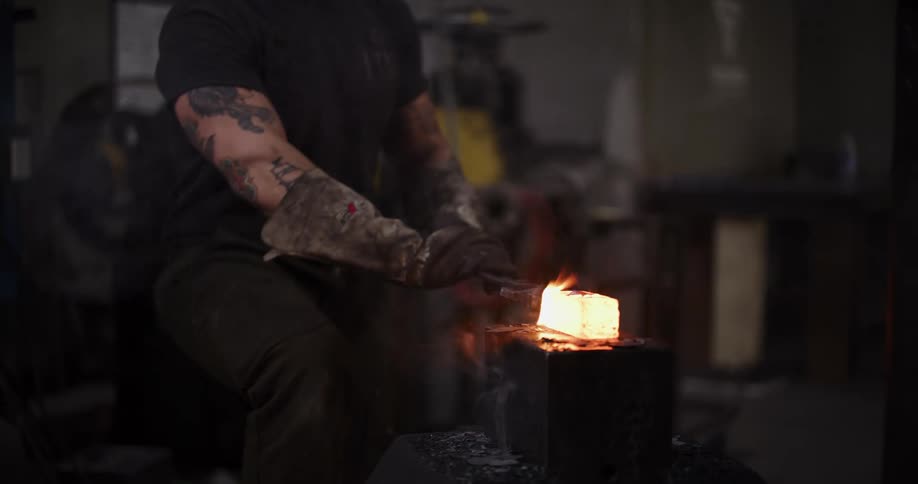 A tattooed blacksmith shapes a glowing hot piece of metal with a powerful industrial hammer in a dark workshop
