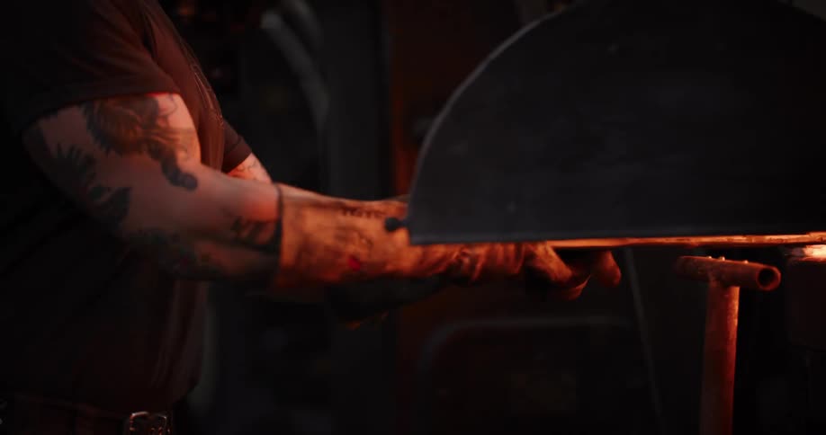 A blacksmith with tattooed arms uses tongs to remove a glowing red-hot piece of metal from a forge in a dark workshop