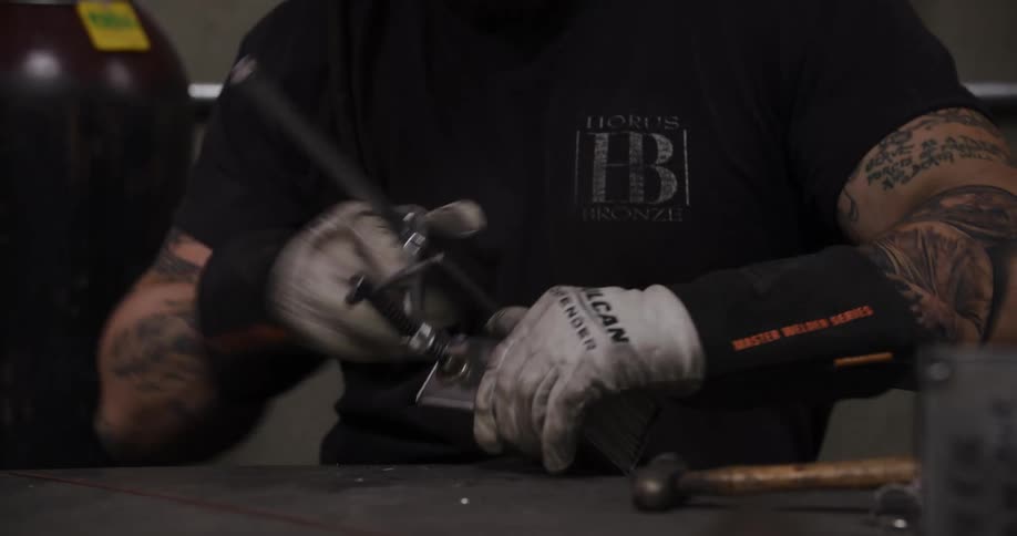 A tattooed metalworker in protective gear uses clamps and a hammer to prepare metal pieces for welding in his workshop