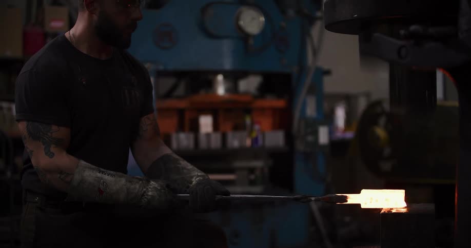 A tattooed blacksmith uses an industrial power hammer to forge a glowing hot piece of metal, sending sparks flying with each impact