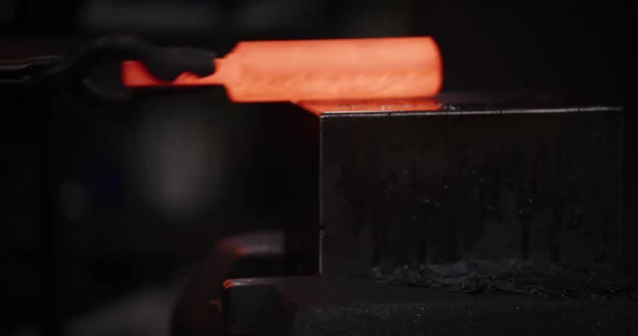 Close-up of a blacksmith holding a glowing hot piece of steel on an anvil as flames erupt in a dark workshop