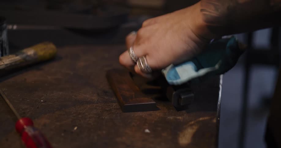 An artisan with rings and tattoos carefully polishes the wooden handle and sheath of a blade in a dimly lit workshop
