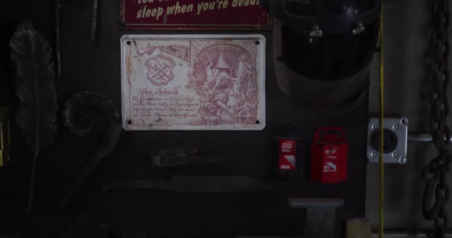A slow vertical pan reveals a blacksmith's workshop, featuring vintage signs, a welding mask, and a cluttered workbench filled with tools and knife blanks