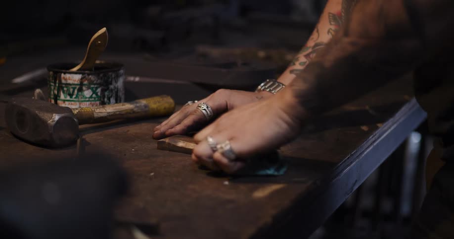 A tattooed craftsman meticulously waxes and polishes a wooden knife handle on a rustic workbench in his workshop